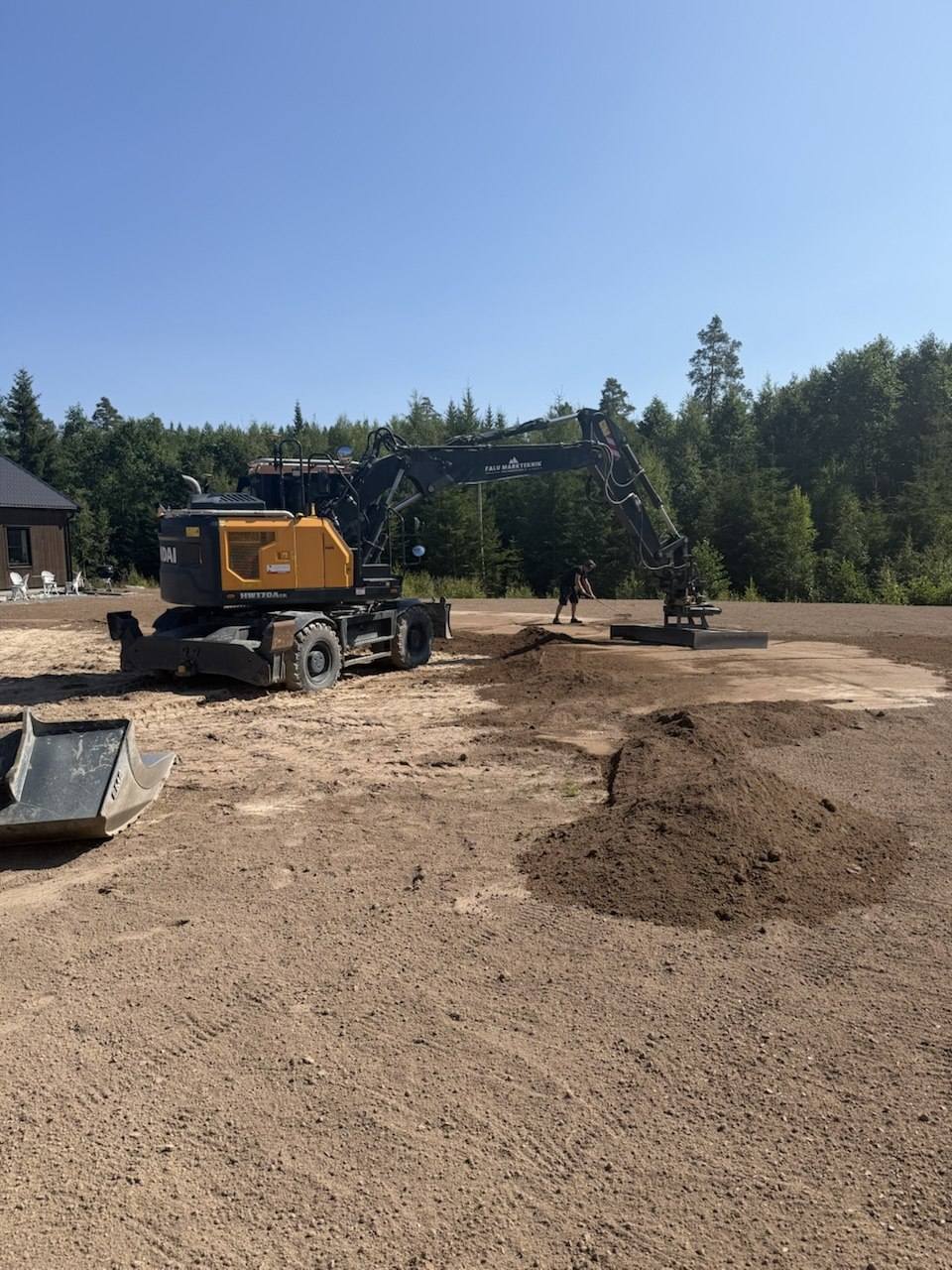 Grävmaskin som jämnar av en tomt med planeringsskopa medan matjord räfsas ut
